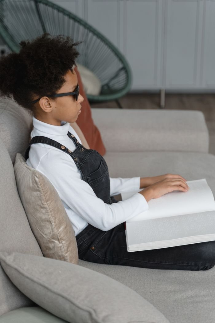 Services A young boy with sunglasses reads braille in a comfortable indoor setting.