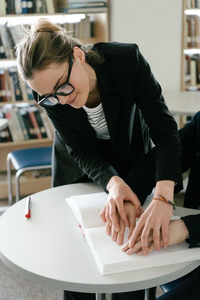 Services Woman guiding a person to read braille in a library, emphasizing support and accessibility.