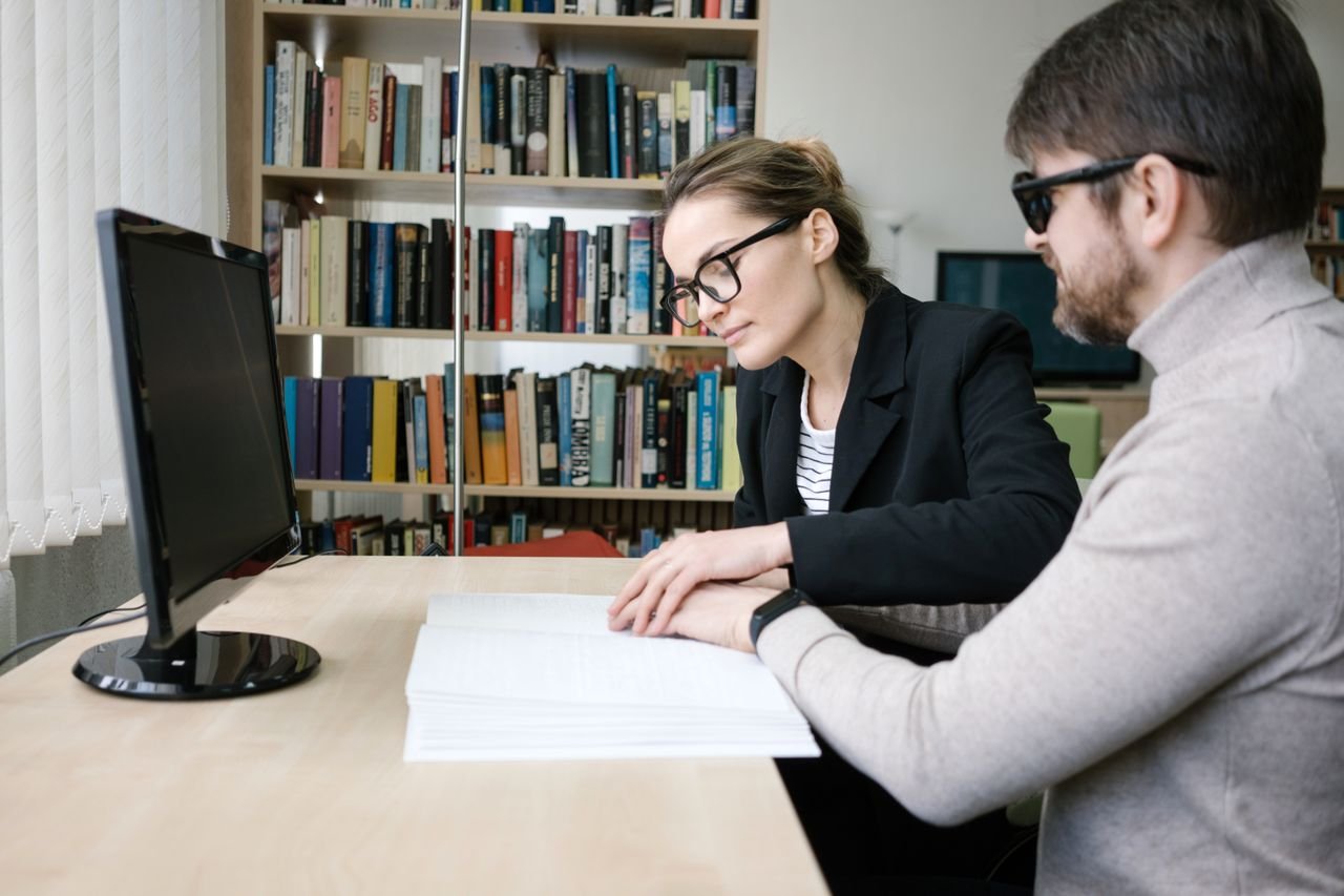 Services A caregiver helps a visually impaired man read a braille book in a library setting.